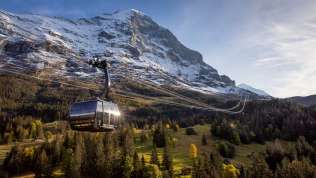 Le téléphérique de l'Eiger Express au printemps, avec un paysage verdoyant au premier plan, des montagnes enneigées en arrière-plan et un ciel bleu.